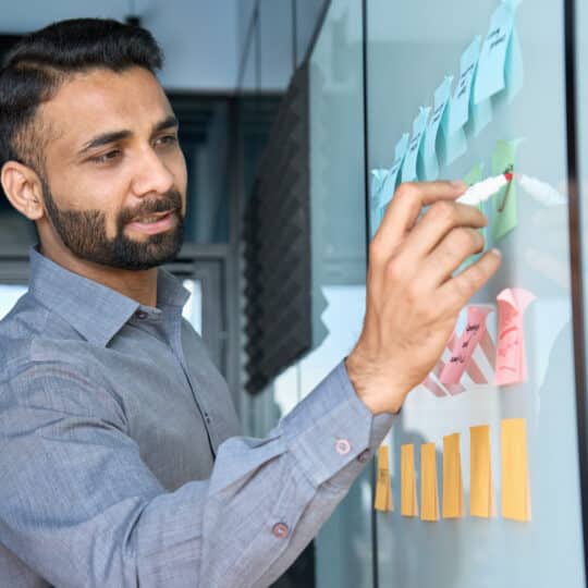 Young indian latin executive manager businessman ceo writing his strategy ideas on sticky notes on whiteboard managing professional organization business plan project presentation in corporate office.